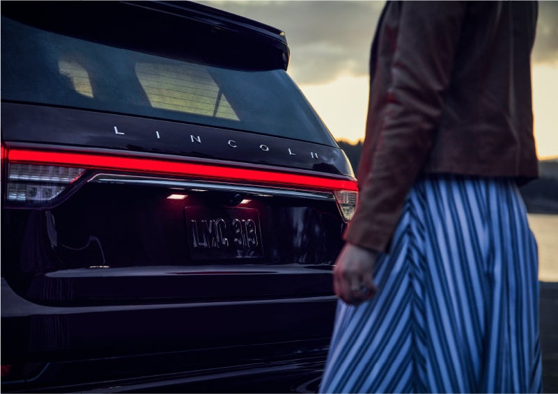 A person is shown near the rear of a 2023 Lincoln Aviator® SUV as the Lincoln Embrace illuminates the rear lights | Karl Malone Lincoln in El Dorado AR
